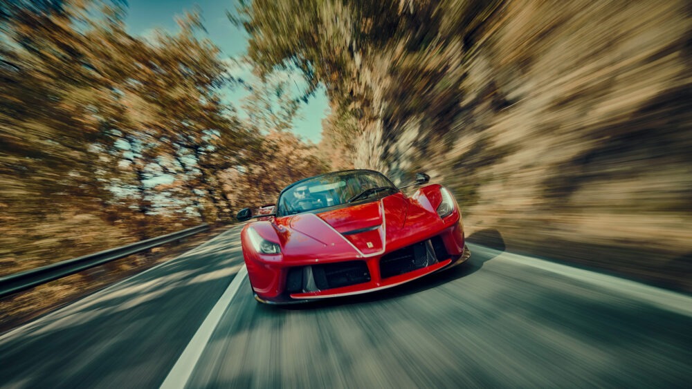 A red sports car speeds along a winding road through a forest. The blurred motion effect highlights the cars velocity, with autumn-colored trees in the background.