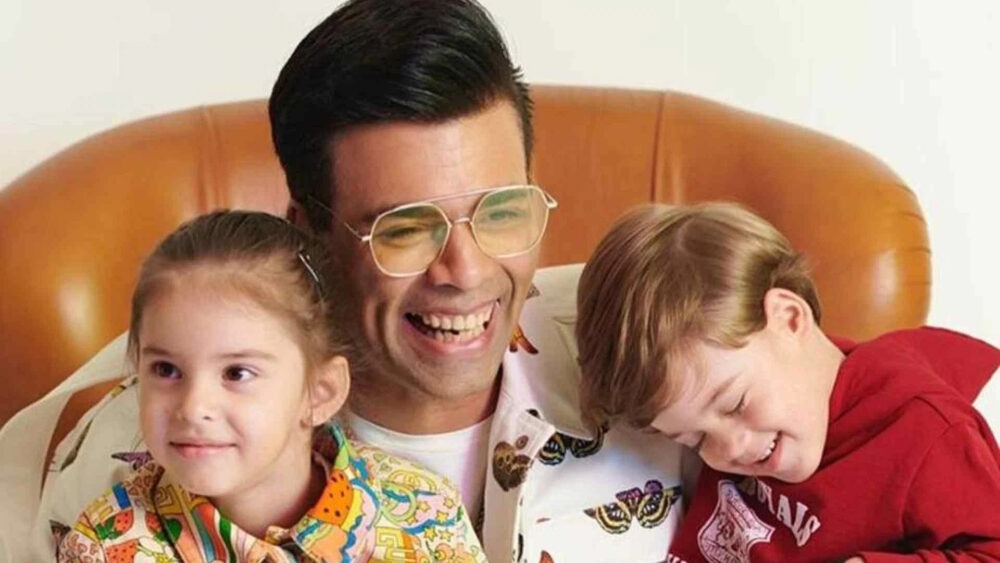 A man resembling Karan Johar, wearing glasses, smiles warmly while sitting on a brown couch with two young children. One child wears a colorful outfit and the other a red shirt, both appearing joyful and relaxed next to him.