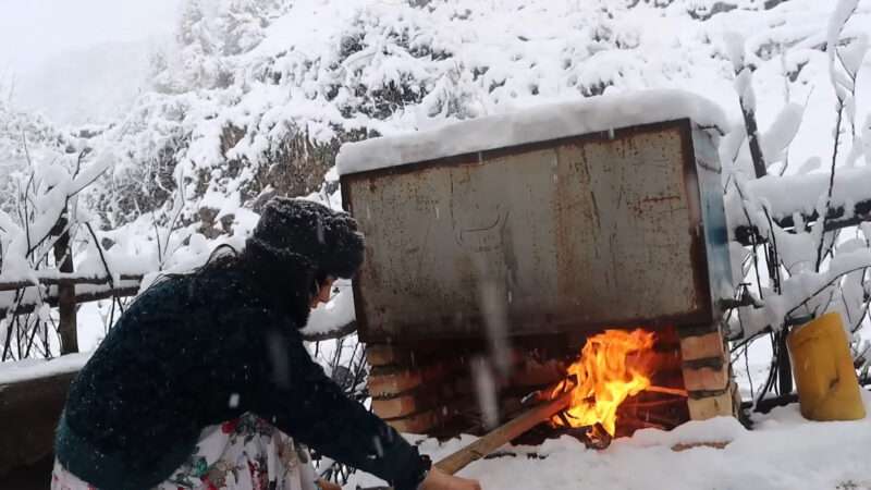 In a snowy setting, a woman feeds wood into a fire to hot water in winter.