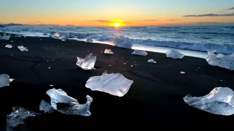 Sunset casts a golden glow on icebergs scattered along a tranquil beach.